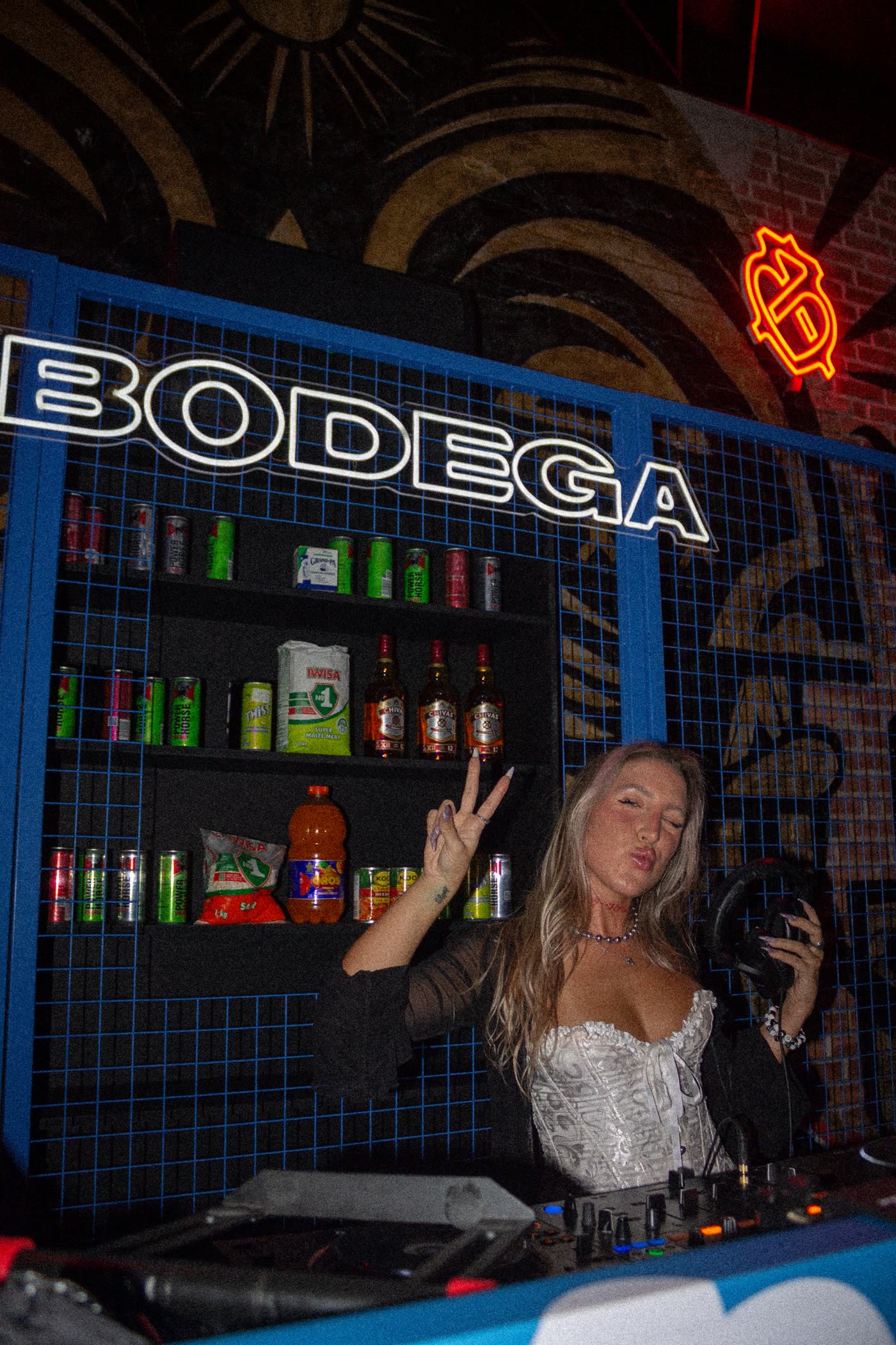 DJ in a white corset with headphones, blowing a kiss under the Bodega neon sign