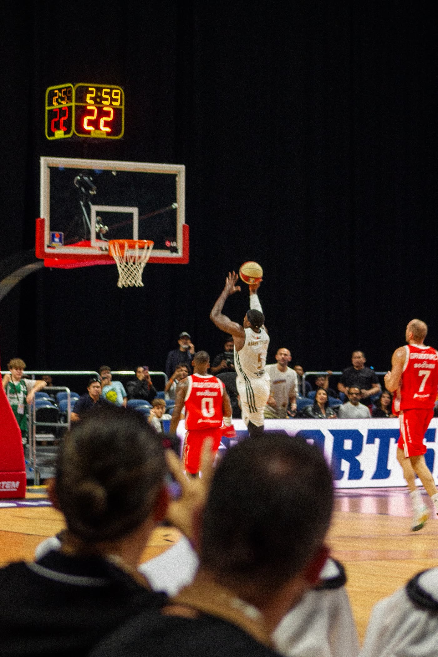 Dubai Basketball player rising for a shot under the shot clock, seen from behind the crowd