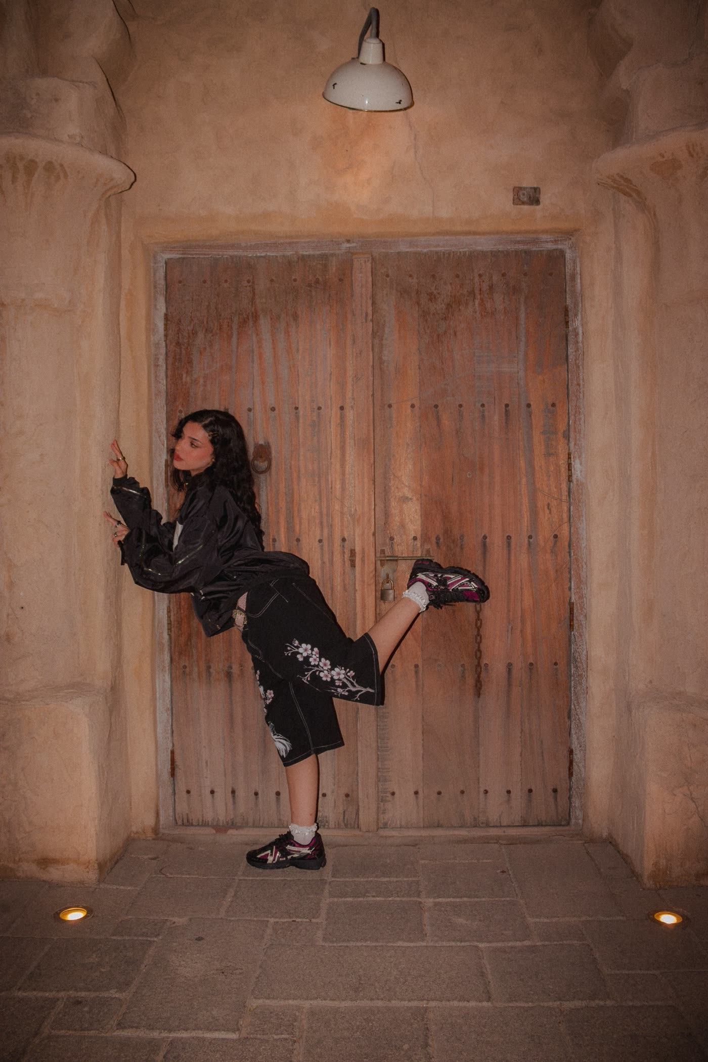 Woman in a black bomber kicking one leg up in front of an old wooden door