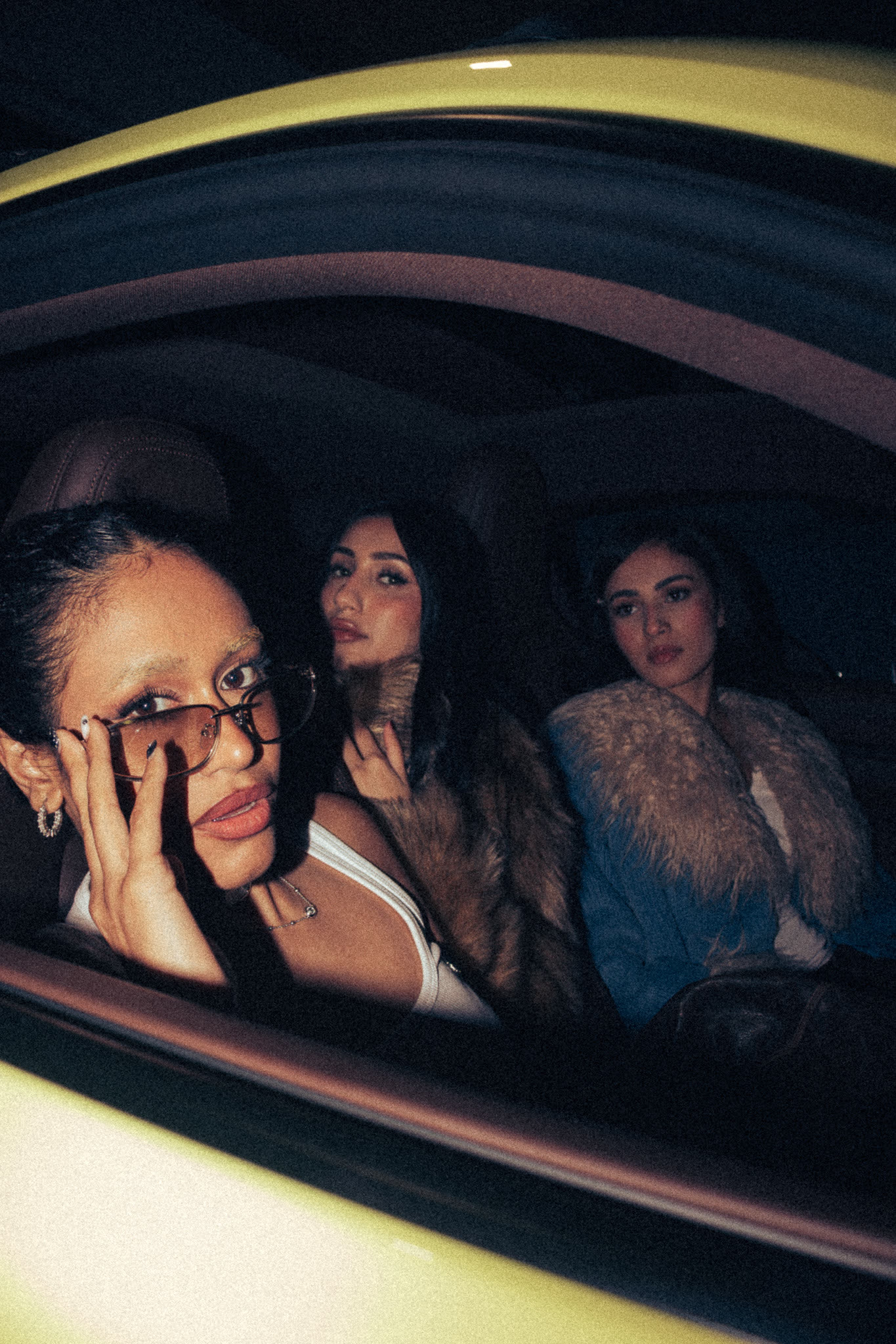 Three women in fur coats in the back of a yellow Porsche, one lowering her sunglasses