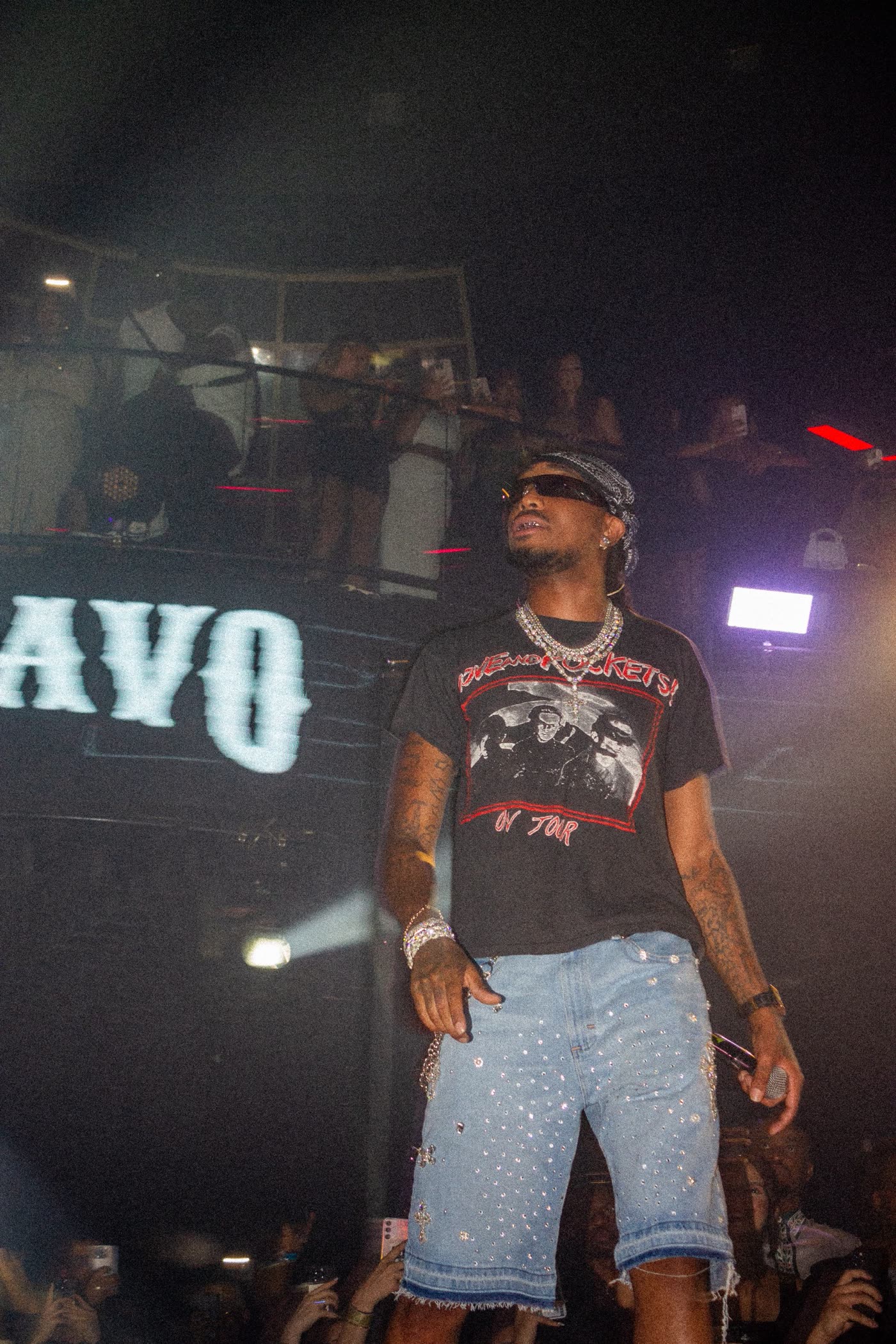 Quavo on stage in sunglasses, chains and crystal denim shorts in front of his name on the screen