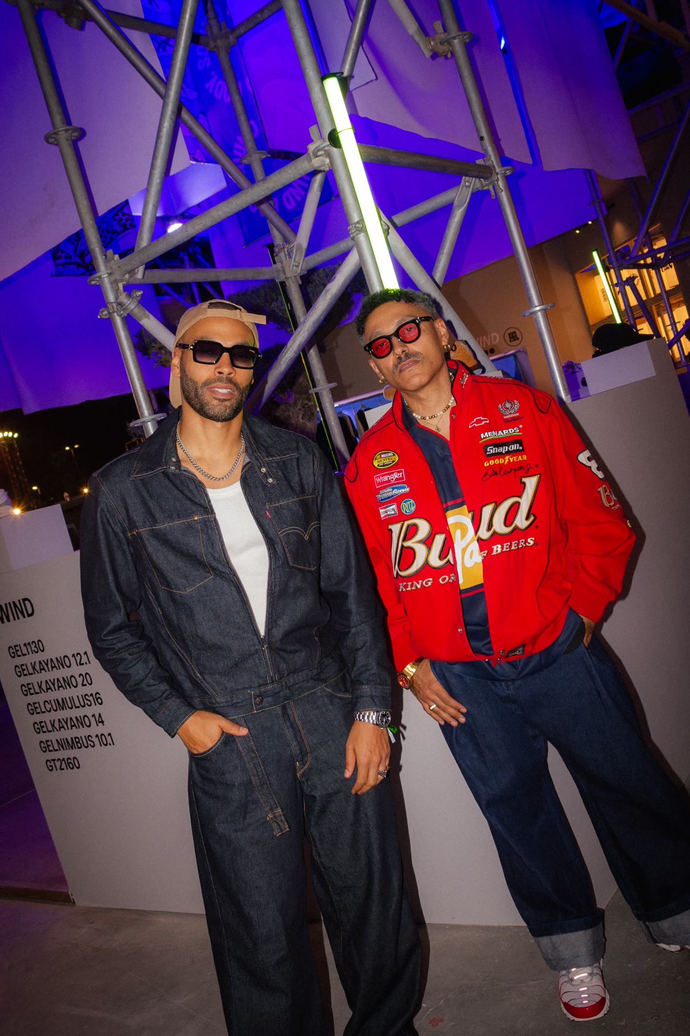 Two men in sunglasses, one in raw denim and one in a red racing jacket, in front of lit scaffolding
