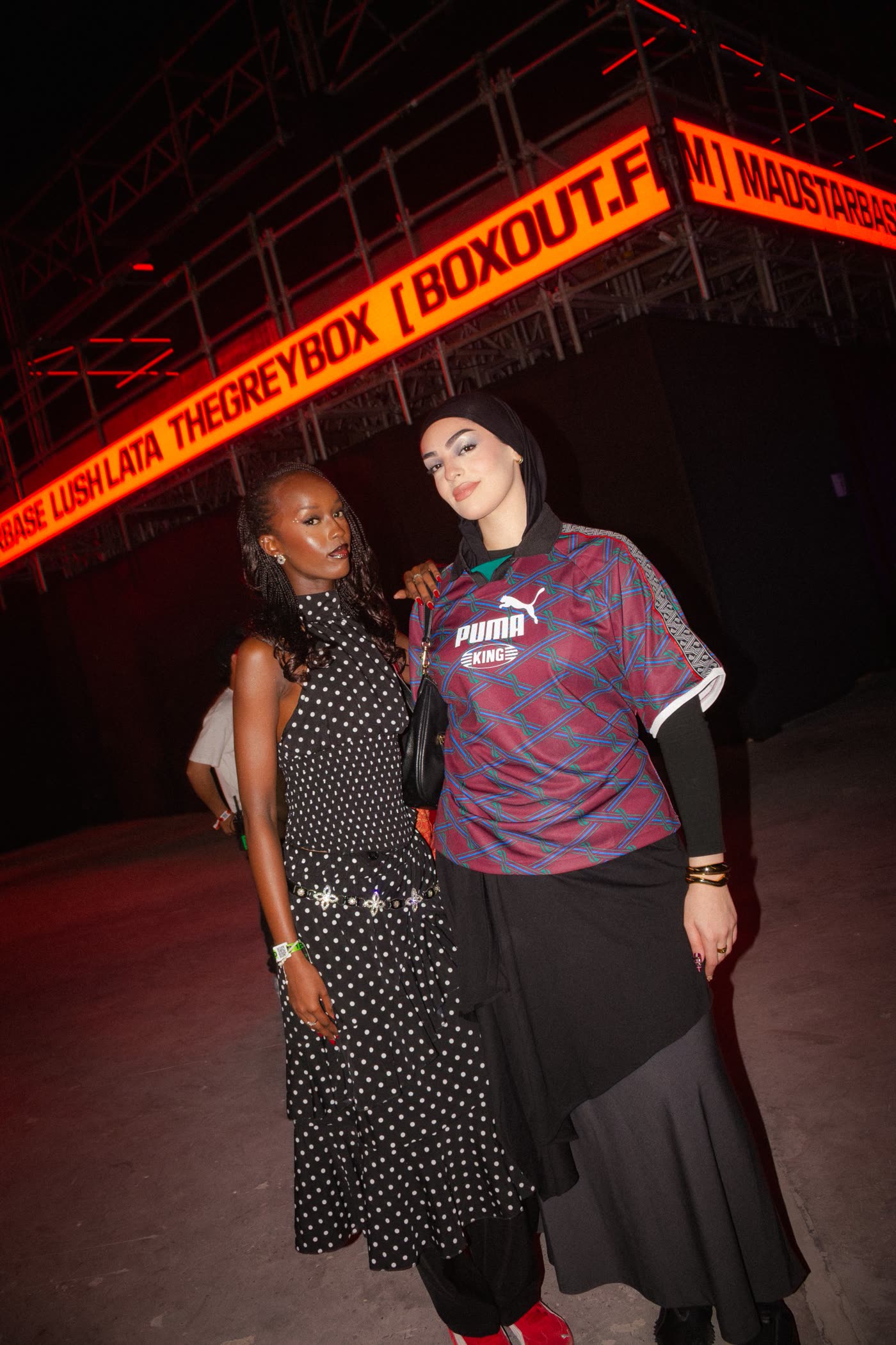 Two women posing under the orange Boxout.fm sign, one in polka dots and one in a Puma King jersey