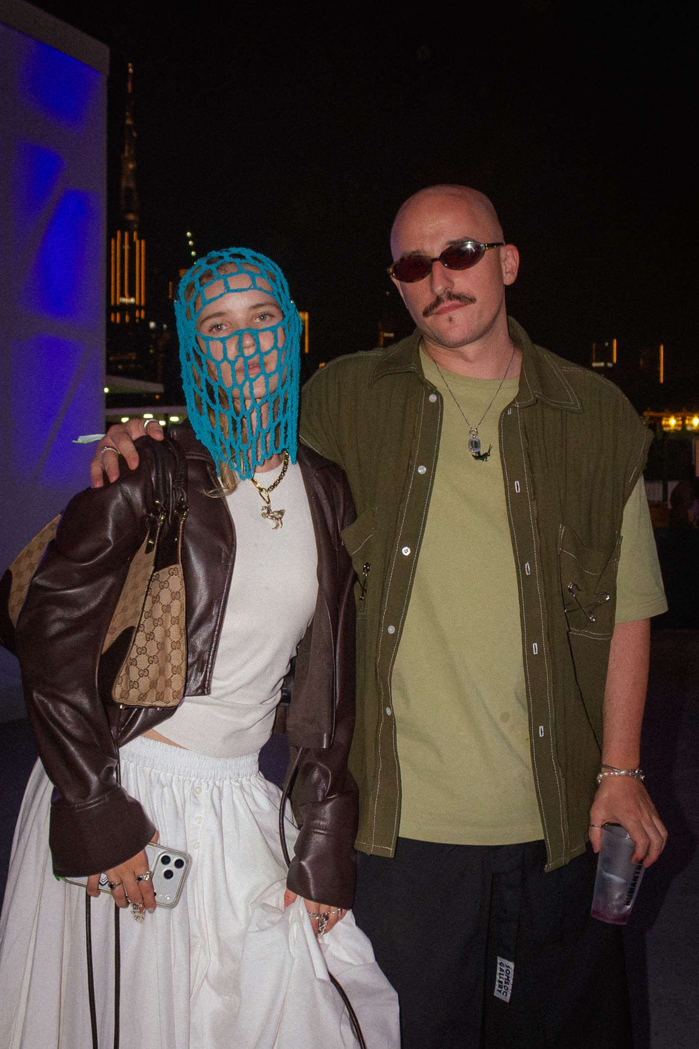 Woman in a blue crochet face mask and leather jacket beside a man in olive with the Dubai skyline behind