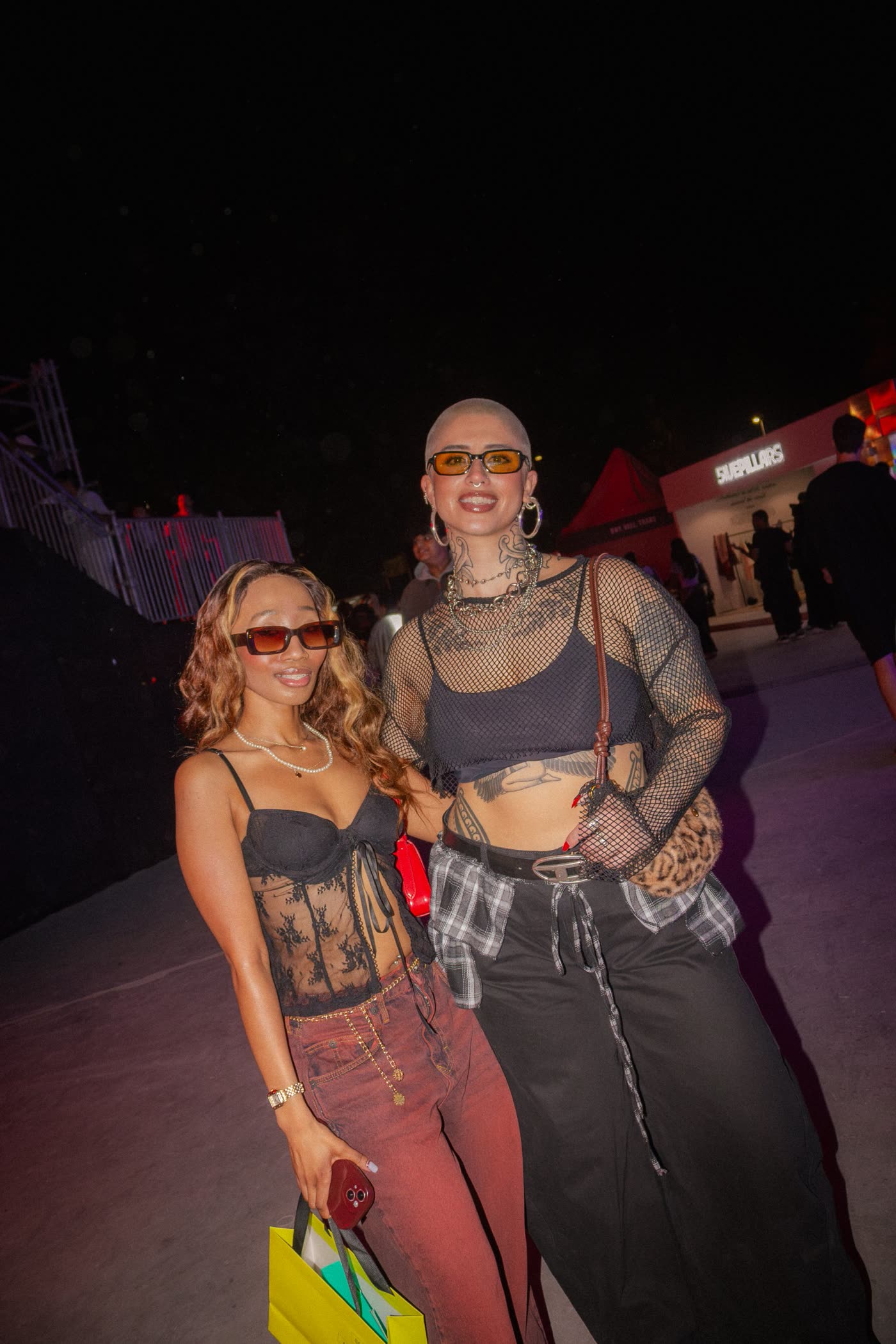 Two friends in lace, mesh and tattoos smiling at the camera at night