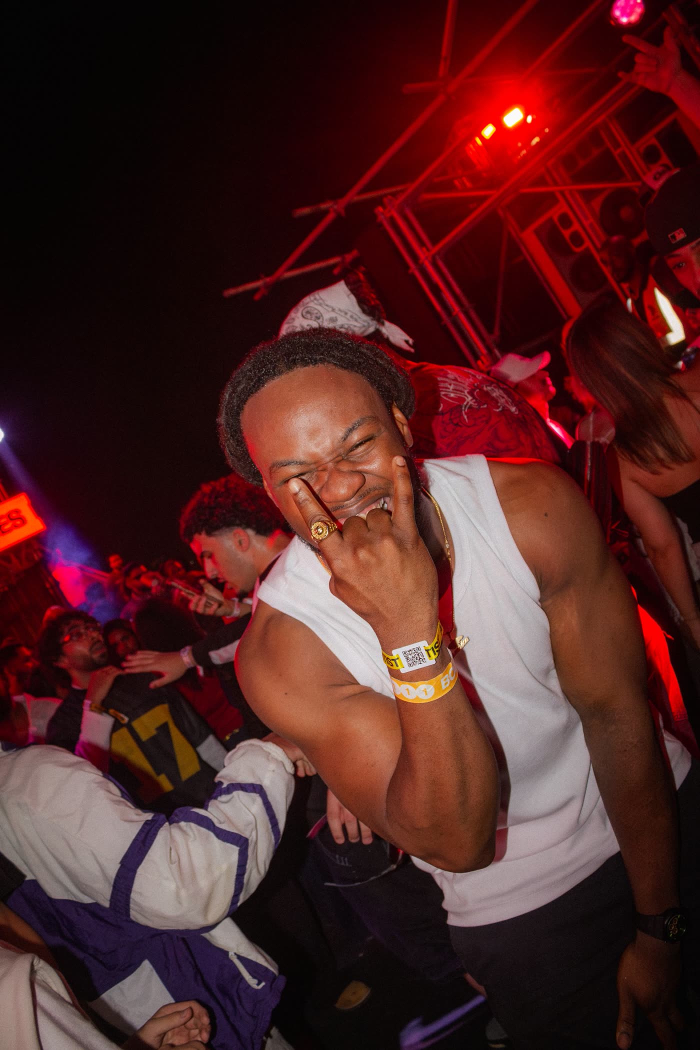 Man in a white vest grinning and pointing at his eye in the red-lit crowd