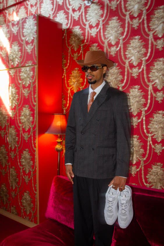 Man in a suit and hat holding sneakers against red wallpaper