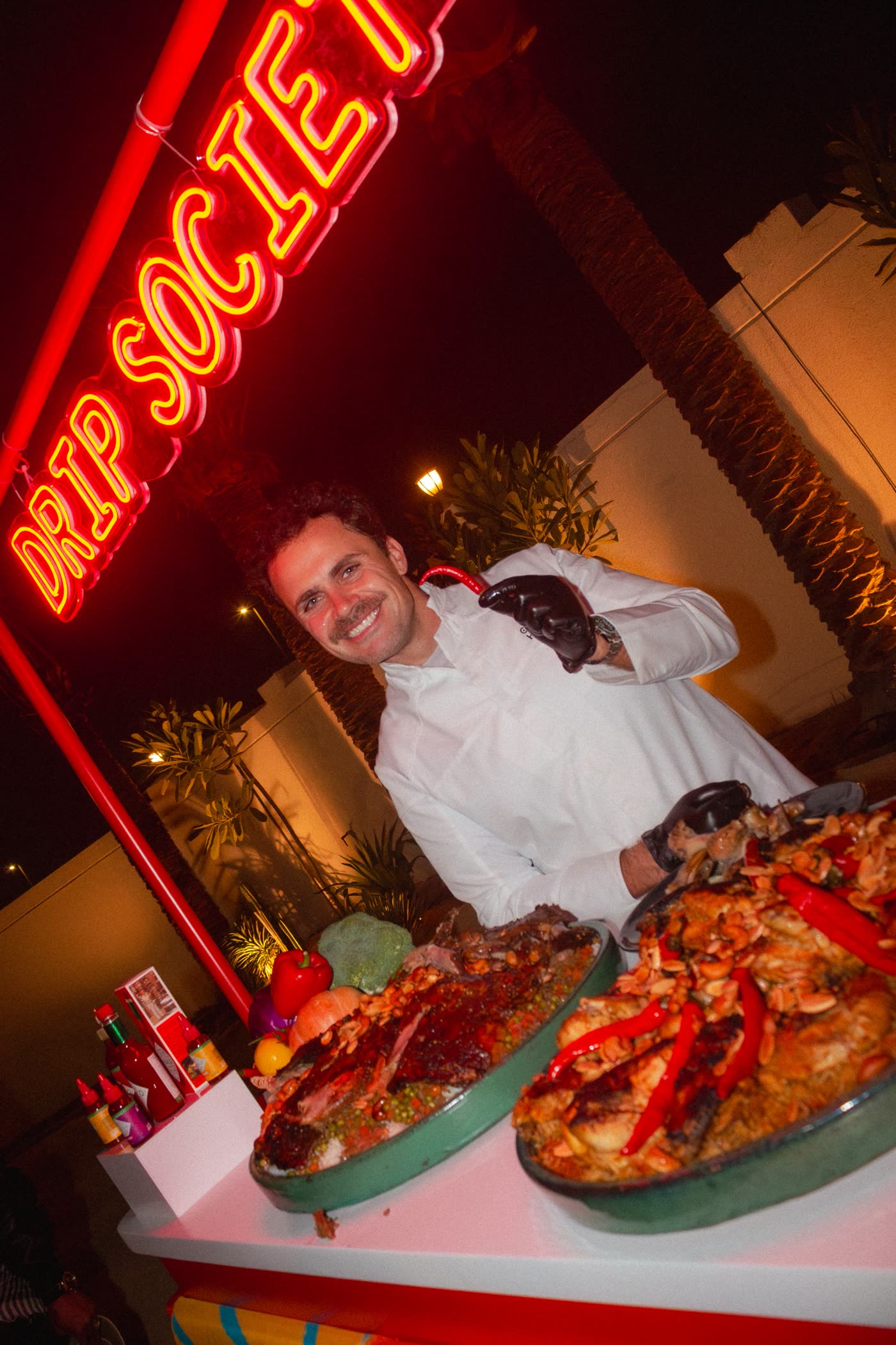 Chef Halawa smiling behind two loaded platters under the red Drip Society neon