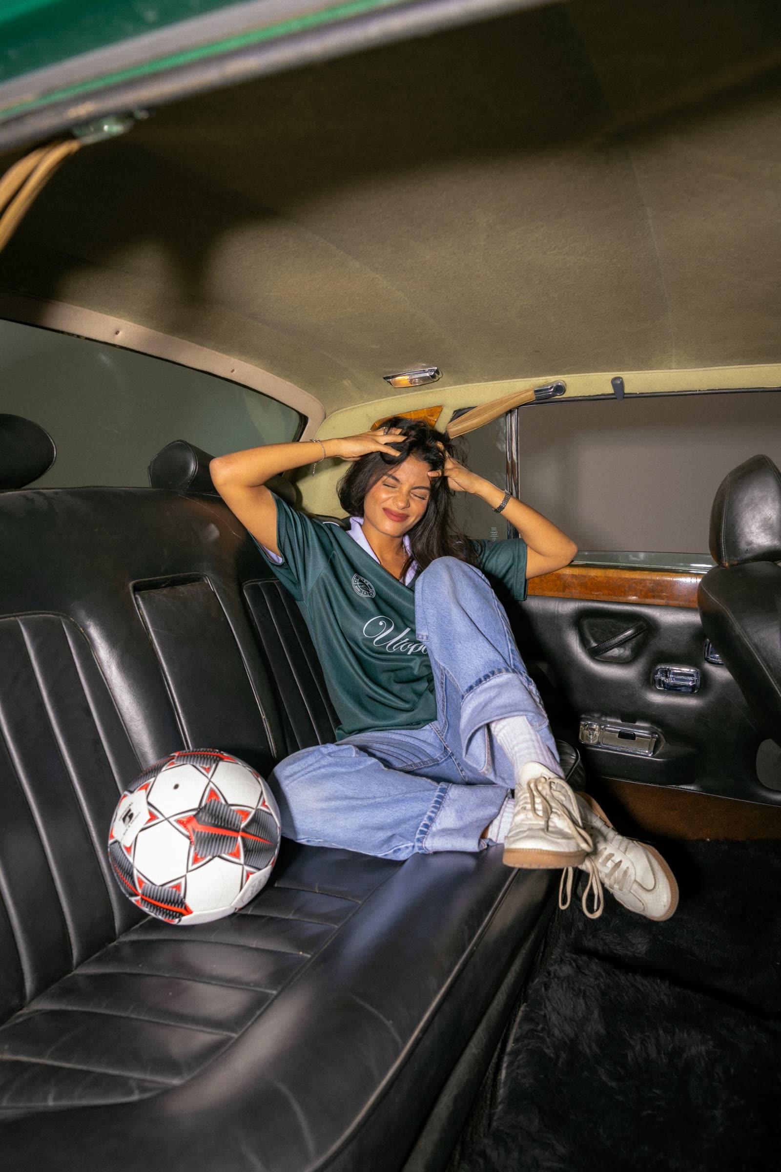 Model in a green Utopia jersey on the back seat of a vintage car with a football