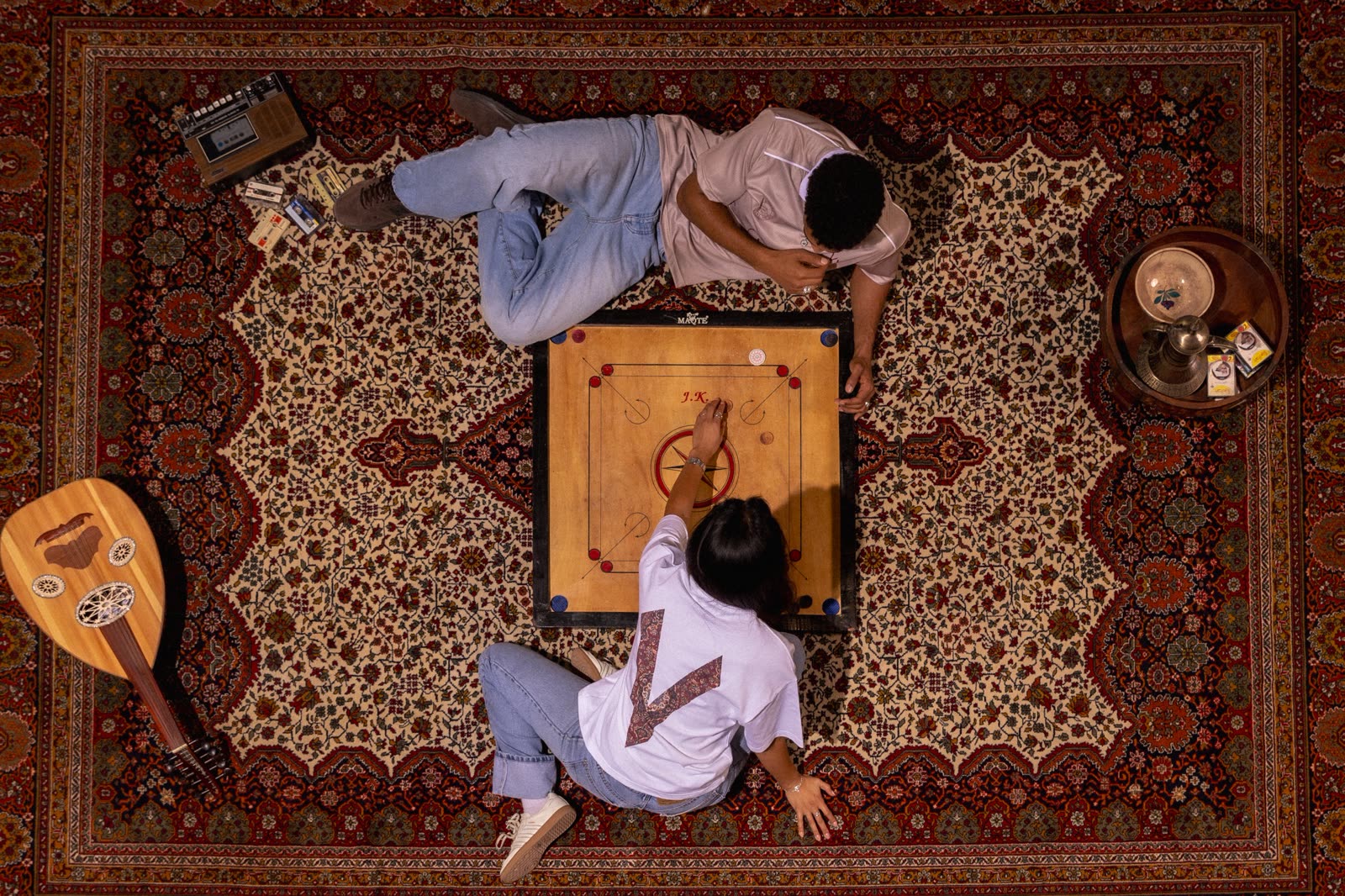 Top-down shot of two models playing carrom on a Persian rug beside an oud and a cassette player